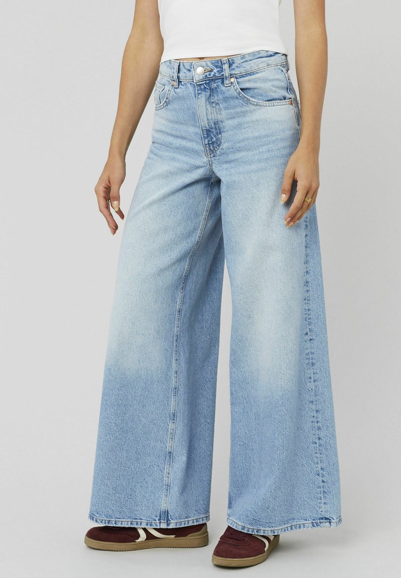 Femme portant un jean large bleu clair, un haut sans manches blanc et des baskets bordeaux, se tenant avec les bras détendus le long du corps.