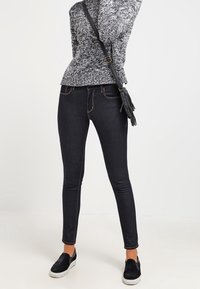 Jeans neri slim-fit con cuciture a contrasto, abbinati a un maglione grigio texturizzato e scarpe nere slip-on, su uno sfondo bianco.