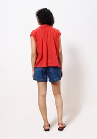 Persona con el cabello rizado que lleva una blusa roja de tirantes suelta, pantalones cortos de denim azules y sandalias rojas, de pie frente a una pared blanca.