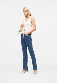 Γυναίκα με λευκό αμάνικο τοπ, μπλε τζιν, μπεζ ψηλοτάκουνα και κρατώντας τσάντα ανοιχτοπράσινου χρώματος στον ώμο μπροστά σε λευκό φόντο.