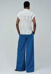 Camisa blanca de manga corta con bordado floral en la parte posterior, combinada con vaqueros de denim azul de pierna ancha. Diseño minimalista, ajuste relajado.