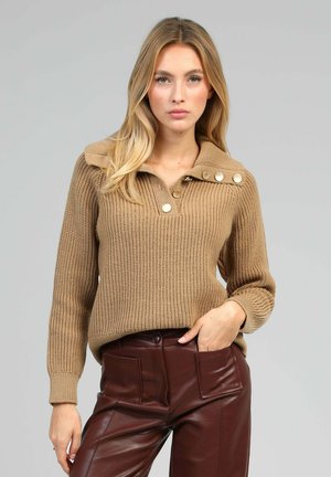 Pull en tricot beige avec un large col, trois boutons au niveau du décolleté, et des poignets côtelés. Assorti à un pantalon en cuir marron foncé.