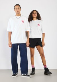 White oversized t-shirts with pink graphics, blue wide-leg jeans, and black boots. Curly hair and braided styles. Plain background.