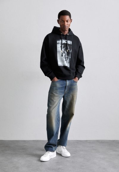 Sudadera negra con estampado gráfico y detalles con cordón, combinada con jeans azul deslavado y zapatillas blancas. El modelo está de pie frente a un fondo neutro.