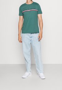 Camiseta verde de manga corta con el logo "HILFIGER" y una franja roja y blanca en el pecho, combinada con jeans de mezclilla azul claro y zapatillas blancas.