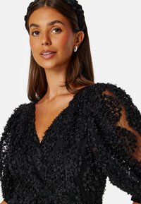 Svart klänning med texturerade blommor, djup V-ringning och genomskinliga puffärmar. Modell bär en svart hårband och stora hoop-örhängen.