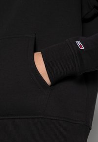 Felpa nera con cappuccio realizzata in cotone spesso, dotata di tasca a canguro e polsini a costine. Piccolo logo ricamato in rosso, bianco e blu sul polsino.