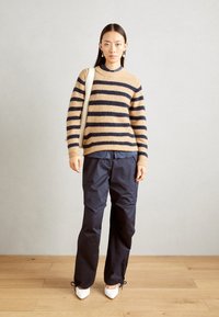 Pulover bej cu dungi bleumarin, cu mansete canelate, asortat cu pantaloni largi bleumarin și pantofi cu vârful ascuțit albi. Modelul poartă o geantă bej.