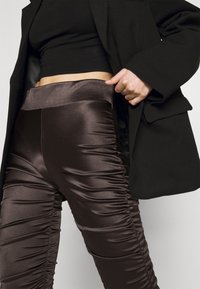 Pantalon taille haute en satin marron avec une texture froncée sur les côtés, associé à un haut noir et une veste noire, mettant en valeur un matériau lisse.