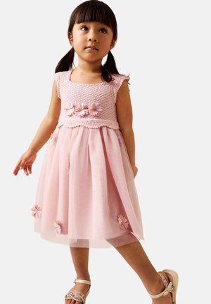 Ung pige i en lyserød ærmeløs kjole med blomsterdetaljer, iført sandaler, stående mod en ensartet lys baggrund.