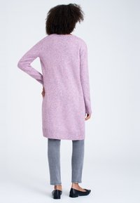 Lång, rosa kofta med en fläckig textur, ribbade ärmslut och nederkant, tillsammans med grå byxor och svarta platta skor, sedd bakifrån.