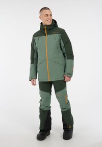 Isolierte Outdoorjacke und -hose in Grün mit dunkelgrünen Akzenten. Verfügt über eine verstellbare Kapuze, Reißverschlusstaschen und eine strapazierfähige, strukturierte Oberfläche.