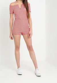 Röd och vit randig off-the-shoulder jumpsuit med V-ringning, åtsittande midja och utsvängda ben, tillsammans med vita sneakers. Slät textur.