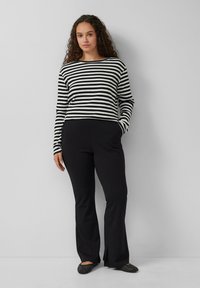 Gestreiftes Langarmshirt in Schwarz und Weiß, kombiniert mit schwarzen Hosen. Das Material wirkt weich, mit einer lockeren Passform und ausgestelltem Bein-Design.