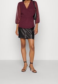 Blusa avvolgente borgogna con maniche trasparenti a pois, abbinata a una mini gonna in pelle nera e tacchi neri con cinturini. Borsa nera semplice.