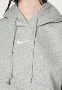 Osoba ve světlém šedém mikinovém svetru s kapucí, na kterém je bílé logo Nike a tkaničky uvázané do uzlů.