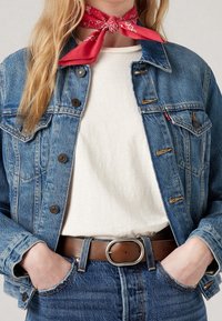 Denimtakki nappilukituksilla, vaalea sininen väri, klassinen malli. Päällä valkoinen paita, yhdistettynä ruskeaan nahkavyöhön ja punaiseen bandanaan.