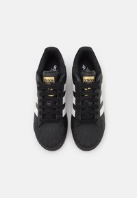 Černé tenisky s gumovou špičkou, třemi bílými pruhy na každé straně a zlatým logem Adidas na jazyku. Šněrovací design, texturovaná látka.