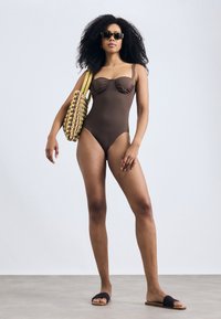 Costume da bagno intero marrone con dettagli arricciati, spalline sottili, abbinato a una borsa a tracolla intrecciata gialla e marrone e sandali scivolosi scuri.