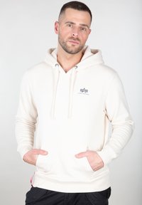 Sudadera blanca con capucha, bolsillos frontales, cordón ajustable y un pequeño logotipo gris en el pecho. Tejido suave y diseño ajustado.