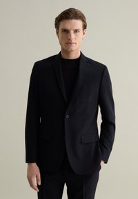 Blazer nero su misura con chiusura a un bottone, revers a punta, due tasche frontali e tessuto liscio. Indossato sopra una camicia scura.