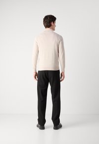 Suéter de punto beige con mangas largas y dobladillo acanalado, combinado con pantalones negros y zapatos oscuros, visto desde atrás.