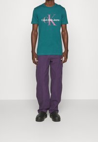 Tyrkysové bavlněné tričko s velkým růžovým a bílým logem "Calvin Klein Jeans", spárované s volnými fialovými cargo kalhotami a černými botami.