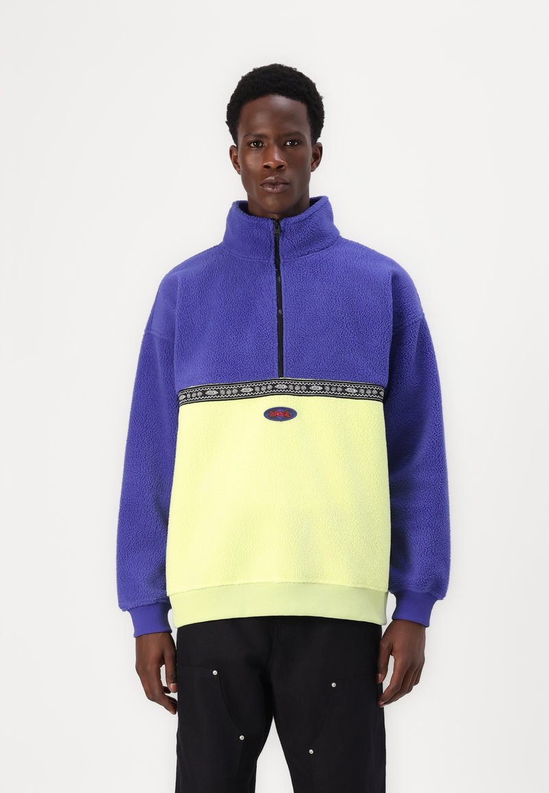 Lila és neon sárga fleece pulóver fél cipzárral elöl, dekoratív fekete csíkkal és bordázott szélekkel. Puha tapintás és laza szabás.