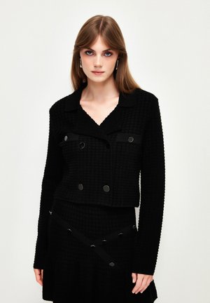 Giacca nera con texture, dal taglio corto, frontale doppiopetto e grandi bottoni decorativi; abbinata a una gonna coordinata con bretelle incrociate.