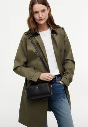 Frau trägt einen olivgrünen Trenchcoat, weißen Pullover, blaue Jeans und eine schwarze Umhängetasche mit goldfarbenem Logo.