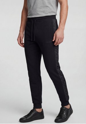 Mand iført sorte joggingbukser med mønstrede side-striber, en grå ærmeløs skjorte og sorte sneakers, der står mod en lys baggrund.