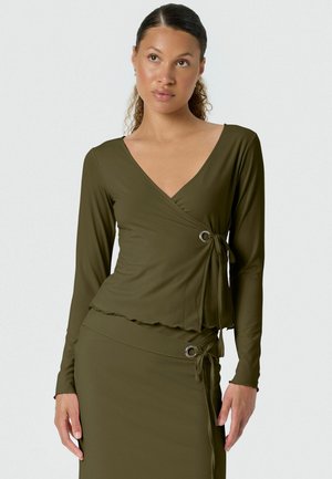 Žena v olivovo zelenom zavinovacom top s dlhým rukávom a ladenej sukni s kovovým kruhom a detailmi na viazanie mašle.