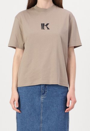 Žena v béžovém tričku s krátkými rukávy a černým logem uprostřed, stojící před jednoduchým pozadím, má na sobě modrou džínovou sukni.