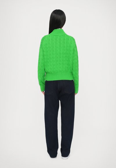 Zelený sveter s vrkočovým vzorom a vysokým golierom, v kombinácii s tmavomodrými voľnými džínsami. Sveter má rebrované manžety a lem.