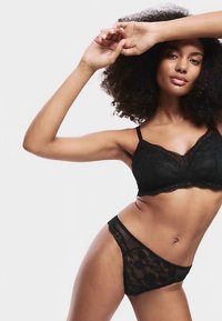 Černý krajkový komplet podprsenky a kalhotek s květinovými vzory, jemnou texturou a vroubkovanými okraji. Modelka pózuje s jednou zvednutou rukou.
