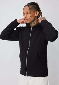 Homme avec des tatouages et des dreadlocks ajustant un sweat à capuche zippé noir, portant un pantalon clair, sur un fond uni clair.