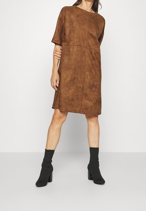 Vrouw die een mid-lengte bruine suède jurk draagt met mouwen tot de elleboog en zwarte enkellaarsjes met hak, staand tegen een witte achtergrond.