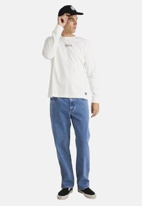 Long-sleeve white t-shirt with black logo, paired with blue denim jeans and black sneakers. The model wears a black cap and poses with one hand raised.