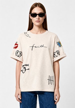 Femme portant des lunettes de soleil, t-shirt beige avec des impressions graphiques incluant le mot foi et jean bleu, debout devant un fond uni.