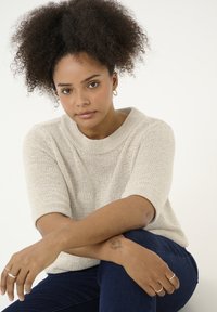 Pull en tricot beige avec un col haut et des manches courtes. Le modèle a les cheveux foncés coiffés de manière volumineuse et porte des bagues. Jeans bleu foncé.