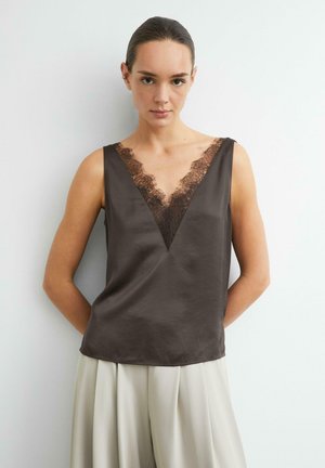 Donna con top in raso marrone scuro senza maniche con rifinitura in pizzo e pantaloni plissettati beige chiaro, in piedi davanti a uno sfondo bianco uniforme.
