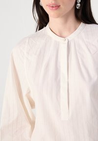 Femme portant un chemisier blanc boutonné avec des rayures verticales fines et un col rond, assorti à de grandes boucles d'oreilles rondes argentées.