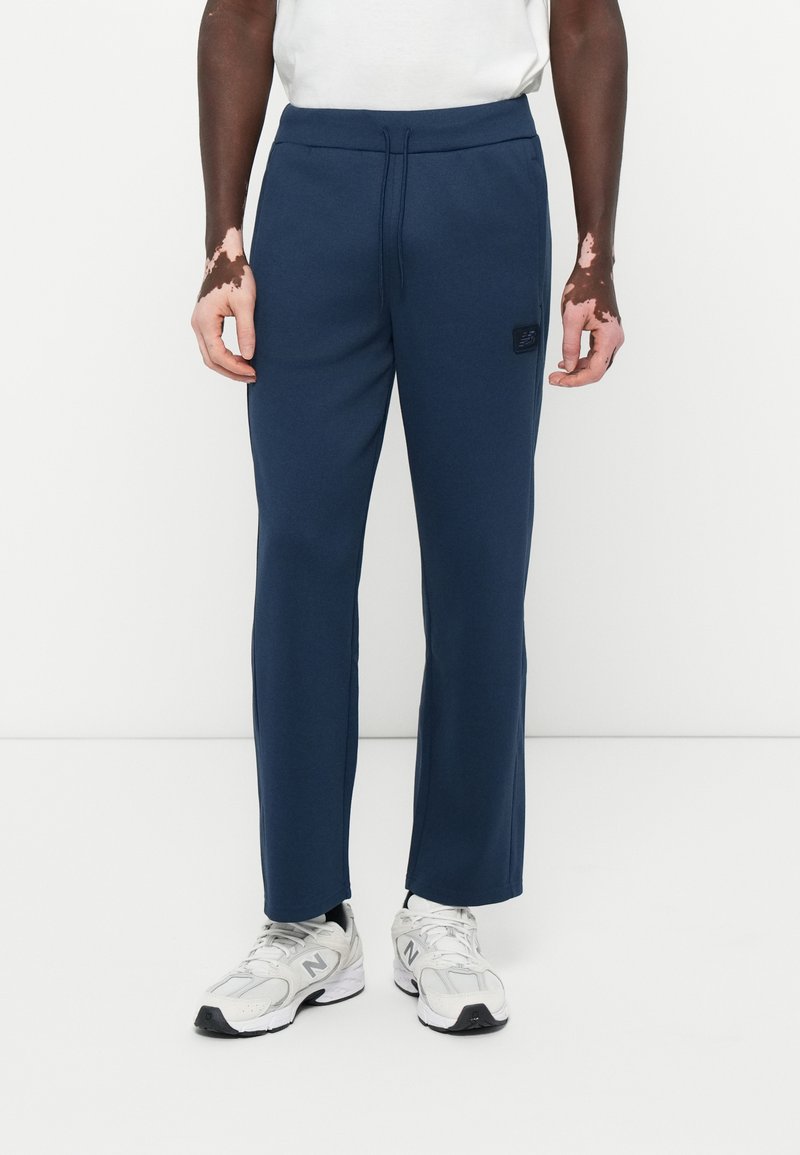 Pantaloni de trening bleu marin cu talie elastică cu șnur, patches cu logo pe o parte și design cu picioare drepte, asociați cu pantofi sport gri deschis.