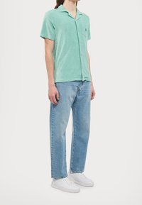 Camicetta verde chiaro a maniche corte con tasca, indossata con jeans azzurri e sneakers bianche. La camicia presenta un design del colletto rilassato.