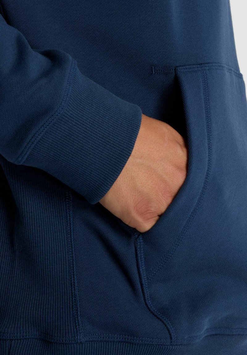 Donkerblauwe sweatshirtstof met geribbelde boorden en een voorvak. De naden zijn zichtbaar langs de randen en het zakje.