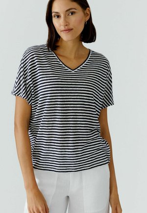 Femme aux cheveux mi-longs portant un haut court à manches courtes à rayures horizontales noires et blanches avec un col en V et un pantalon blanc, debout devant un fond uni.