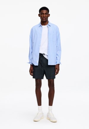 Homme debout portant une chemise à rayures bleu clair boutonnée sur un t-shirt blanc, un short noir, des chaussettes blanches et des baskets blanches, sur fond blanc.
