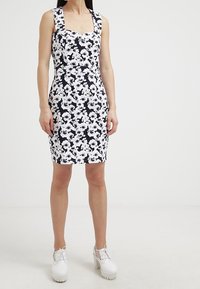 Kvinna iklädd en ärmlös svart och vit blommig figurnära klänning, tillsammans med vita platåsnörskor, stående mot en neutral bakgrund.