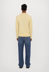 Světle žlutá polo košile s dlouhým rukávem, límečkem a žebrovaným lemem, v kombinaci s volnými modrými džíny z denimu obsahujícími zadní kapsy.