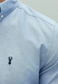 Jasnoniebieska koszula na guziki z fakturowanej tkaniny. Z przodu, przy piersi, niewielkie ciemnoniebieskie logo w kształcie homara. Casualowy krój z klasycznym kołnierzem.